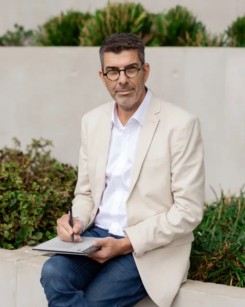 Jonathan Rich, wearing round glasses, seated outdoors on a low wall with a tablet and pen.