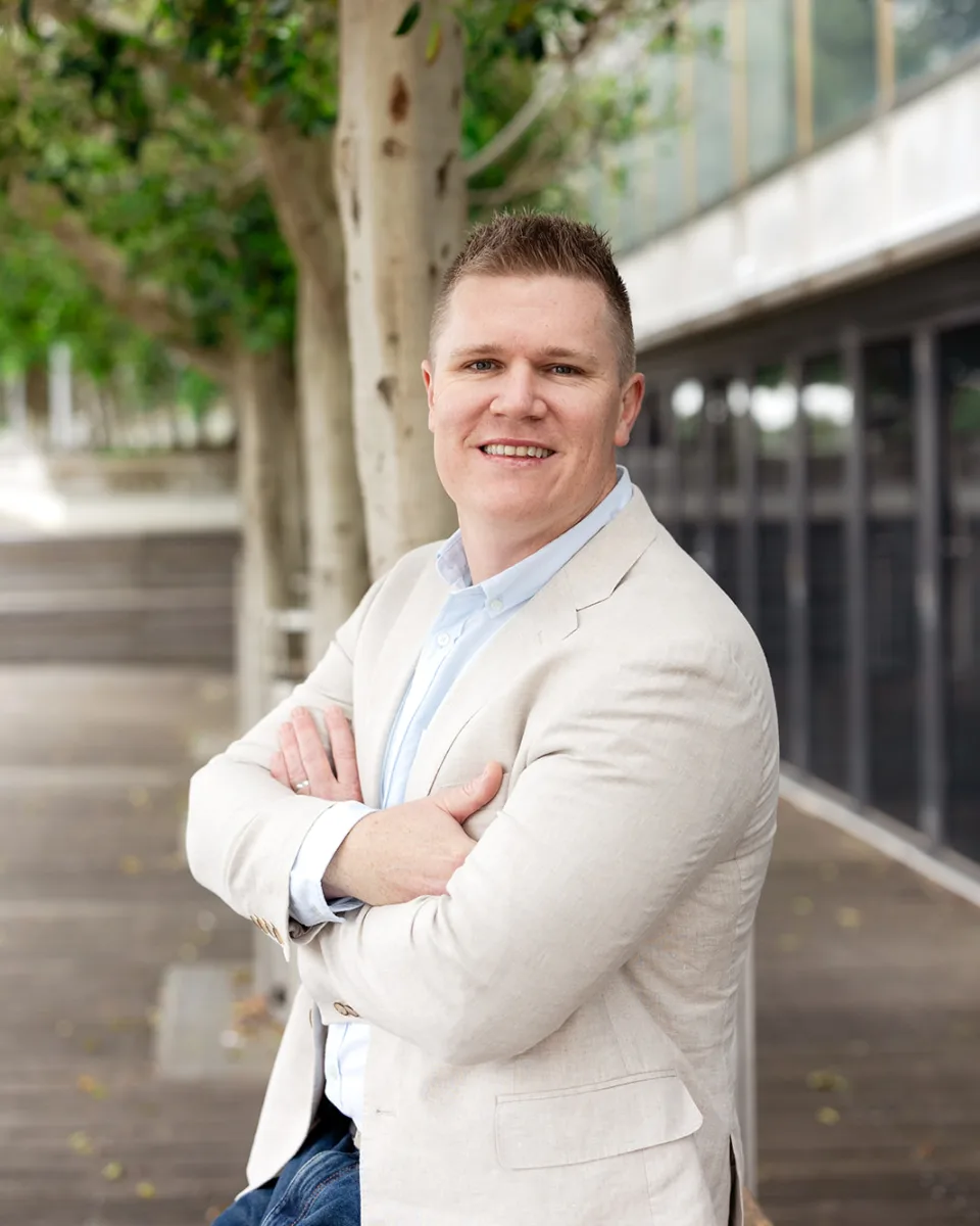 Shane Scriven, smiling with arms folded, standing outdoors beside a row of trees.